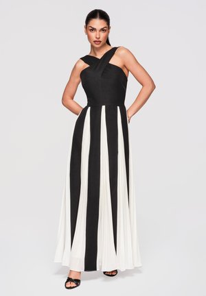 Vrouw staat met haar handen achter haar rug, gekleed in een zwart-witte mouwloze floorlength jurk met verticale strepen en sandalen.