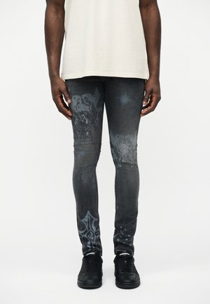 Férfi vékony szabású fekete farmert visel, melyen kopott koponya és kereszt minták vannak, fekete sportcipővel és bézs inggel, fehér háttér előtt áll.