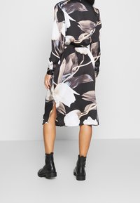 Frau in schwarzen Knöchelboots und einem schwarzen Midikleid mit beigen und weißen großen Blumenmustern, von hinten gesehen.