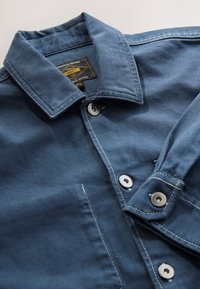 Blå denimjakke med standardkrave, sølvfarvede trykknapper og kontrasterende hvide syninger. Har en label på indersiden af nakken.