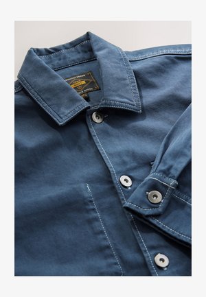 Giacca di jeans blu con colletto standard, bottoni automatici color argento e cuciture a contrasto bianche. Presenta un'etichetta all'interno del collo.