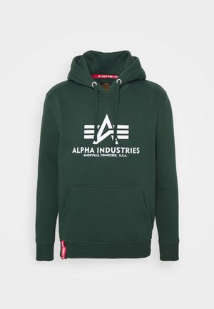 Sötétzöld kapucnis pulóver elülső zsebbel, fehér logóval és "ALPHA INDUSTRIES" felirattal a mellkasán. Húzózsinórral ellátott kapucni és bordázott mandzsetták jellemzik.
