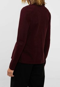 Burgundy långärmad kavaj med en slät textur, figursydd design och kavajkrage, i kombination med svarta byxor.