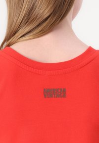 Piros pamut póló lekerekített nyakkivágással, a hátsó részen, a gallérhoz közel, egy kis fekete "AMERICAN VINTAGE" logóval varrva.