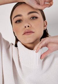 Hellrosa gestrickter Pullover mit hohem, geripptem Kragen; zeichnet sich durch einen lockeren Schnitt und eine weiche Textur aus, teilweise sichtbare Hände, die den Kragen anpassen.