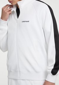 Witte zip-up sweatshirt met zwarte zijpanelen, voorzien van een hoge kraag en geborduurd "GUESS JEANS" logo op de borst. Glad textuur.