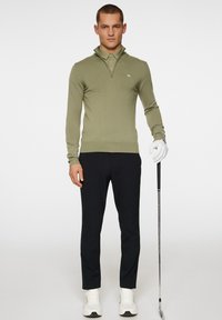 Olivgrön zip-up tröja med hög krage, kroppsnära design och ribbad nederkant, i kombination med svarta slim-fit byxor och vita sneakers.