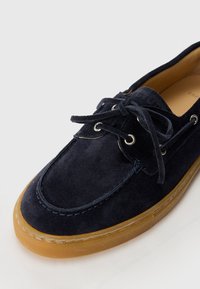 Marineblauer Wildlederbootsschuh mit leichtem Gummisohle, runder Zehenpartie und schnürbarem Design, das mit metallischen Ösen und tonalen Nähdetails versehen ist.