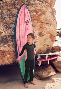 Black wetsuit with pink accents on arms; child holds a multicolored surfboard with pink and green swirls. Rocky beach backdrop.