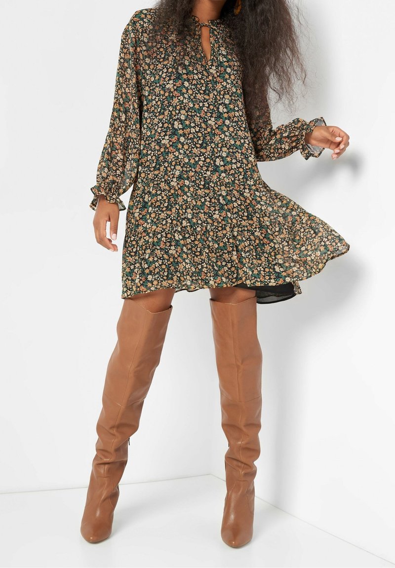 Blommig klänning i mörkgrönt med trumpetärmar och nyckelhålsknäppning. Matchad med bruna läderstövlar som går över knäna.