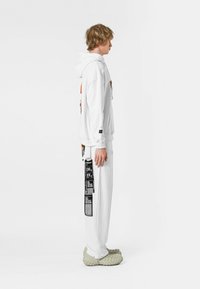 Sudadera blanca con un diseño gráfico frontal, combinada con pantalones blancos que tienen una etiqueta de impresión negra. Usada con calzado texturizado de color gris claro.