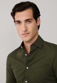 Camisa verde oscura de botones con un cuello estructurado, botones blancos y un sutil detalle de emblema en el lado izquierdo del pecho. Tejido suave y texturizado.