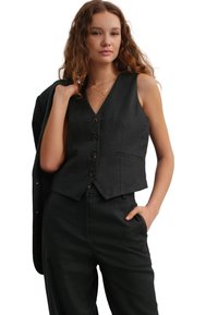 Gilet in lino nero con scollo a V, cinque bottoni marroni e tasca laterale, indossato con pantaloni abbinati. Il modello tiene una giacca in mano.