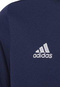 Tumši zila teksturēta auduma, ar baltu izšūtu Adidas logo augšējā kreisajā stūrī. Dizains piedāvā klasisku sporta piegriezumu.