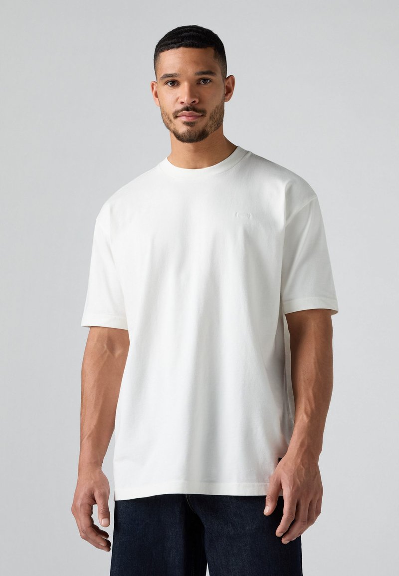 Biała koszulka z krótkim rękawem i okrągłym dekoltem, gładka w dotyku, z delikatnym logo na klatce piersiowej. Połączona z ciemnoniebieskimi dżinsami.