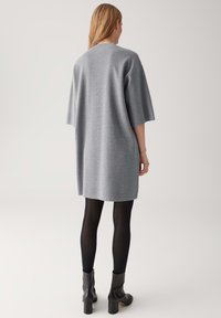 Frau in einem locker sitzenden, grauen knielangen Kleid mit halben Ärmeln, schwarzen Strumpfhosen und schwarzen Knöchelstiefeln, die vor einem schlichten Hintergrund steht.