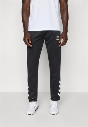 Pantaloni de trening negri cu accente albe în formă de v în părțile laterale, realizați dintr-un material fin. Asortați cu adidași albi și o cămașă deschisă.