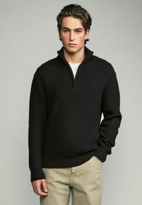 Schwarzer Strickpullover mit hohem Kragen und halbem Reißverschluss. Verfügt über eine gerippte Textur und lange Ärmel, getragen über einem weißen Hemd.
