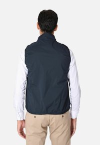 Gilet senza maniche blu navy realizzato in tessuto leggero e lucido con colletto alto. Presenta una superficie liscia e un orlo elastico, indossato sopra una camicia leggera.