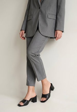 Osoba ubrana w szary garnitur z dopasowanymi spodniami i czarnymi, odkrytymi na palce butami na obcasie typu block, stojąca na jednolitym beżowym tle.