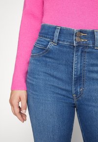Vaqueros de tiro alto de color azul con una textura lisa, que cuentan con dos botones, costuras claras y bolsillos frontales. Combinados con un suéter de canalé rosa.