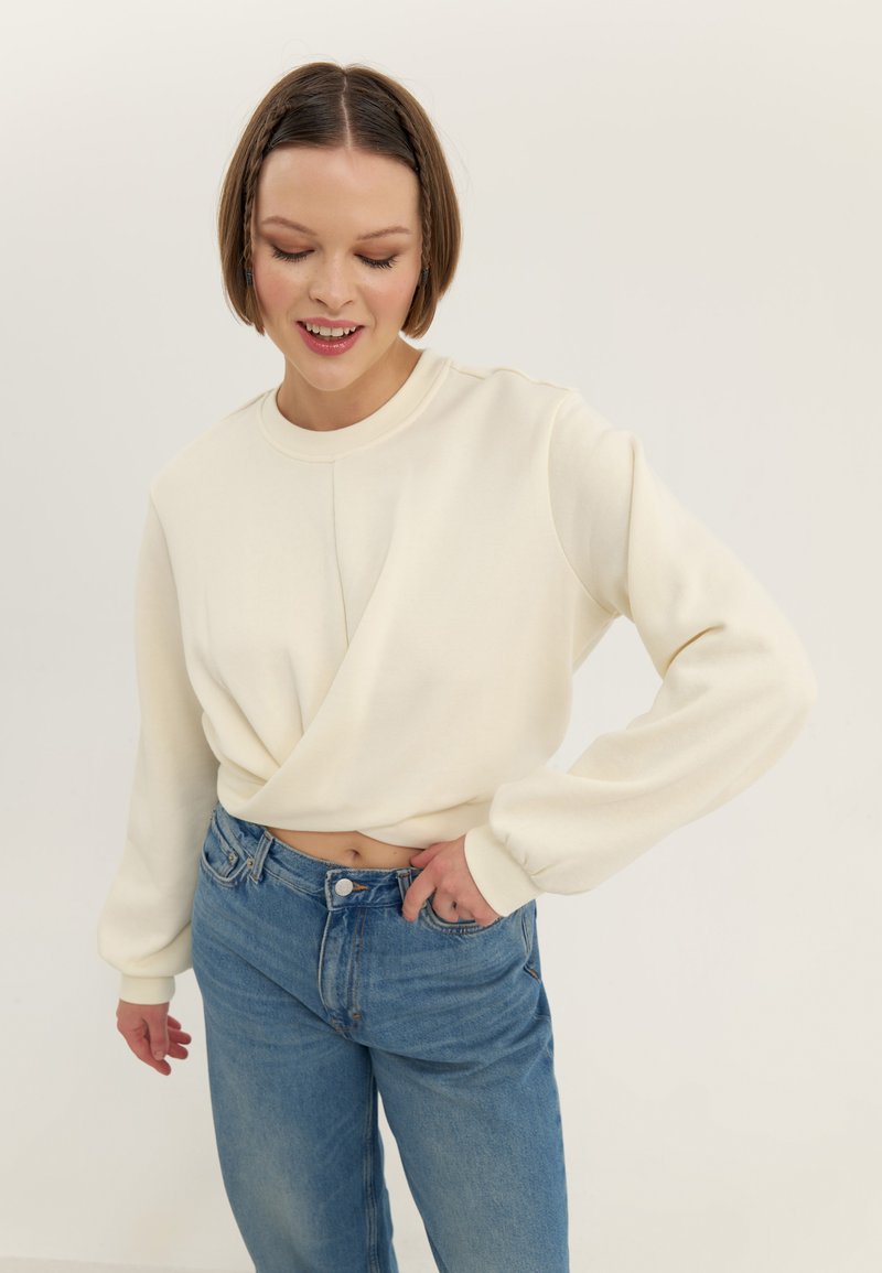 Felpa corta color crema con maniche lunghe, caratterizzata da un design incrociato sul davanti e una texture liscia, abbinata a jeans blu a vita alta.