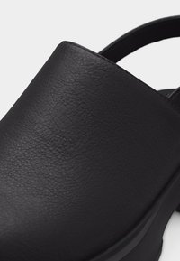 Μαύρο δερμάτινο παπούτσι τύπου slip-on με υφή στην επιφάνεια και χαμηλό τακούνι, με λουράκι στο πίσω μέρος για ασφαλή εφαρμογή.