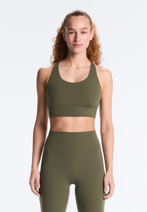 Femeie cu păr creț, purtând un sutien sport de culoare verde măsliniu și colanți cu talie înaltă, stând pe un fundal simplu și deschis la culoare.