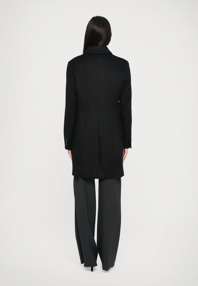 Black wool coat with a notched collar, tailored fit, and rear vent. Paired with loose grey trousers featuring vertical stripes.