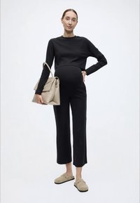 Top preto com mangas compridas caneladas, calças pretas curtas e tamancos bege de camurça. A modelo segura uma mala bege clara com design estruturado.