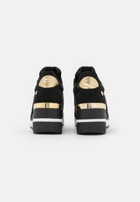 Black suede sneakers with glossy gold accents at the heel, featuring a white stripe on the sole for contrast and a textured upper design.