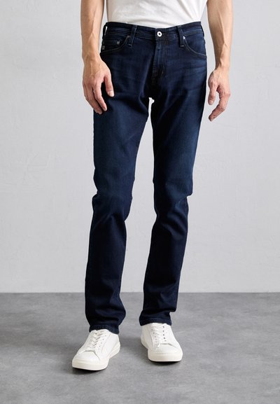 Jeans in denim blu scuro con vestibilità slim, design a cinque tasche e chiusura con bottone metallico, abbinati a scarpe da ginnastica bianche.