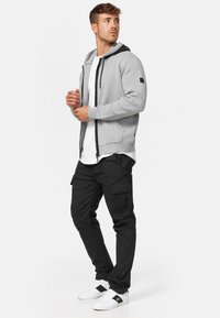 Un sweat à capuche gris avec des accents noirs et une fermeture éclair, associé à un pantalon cargo noir et des baskets blanches. Le modèle se tient dans une position neutre.