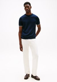 Maglione a maniche corte in maglia blu navy con polsini a righe bianche, abbinato a pantaloni bianchi e mocassini marrone scuro. Design semplice, texture liscia.