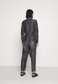 Mono de mezclilla oscura con mangas largas y cintura elástica, que presenta una pretina trasera y puños enrollados. Acentuado con bolsillos laterales y un diseño simple.