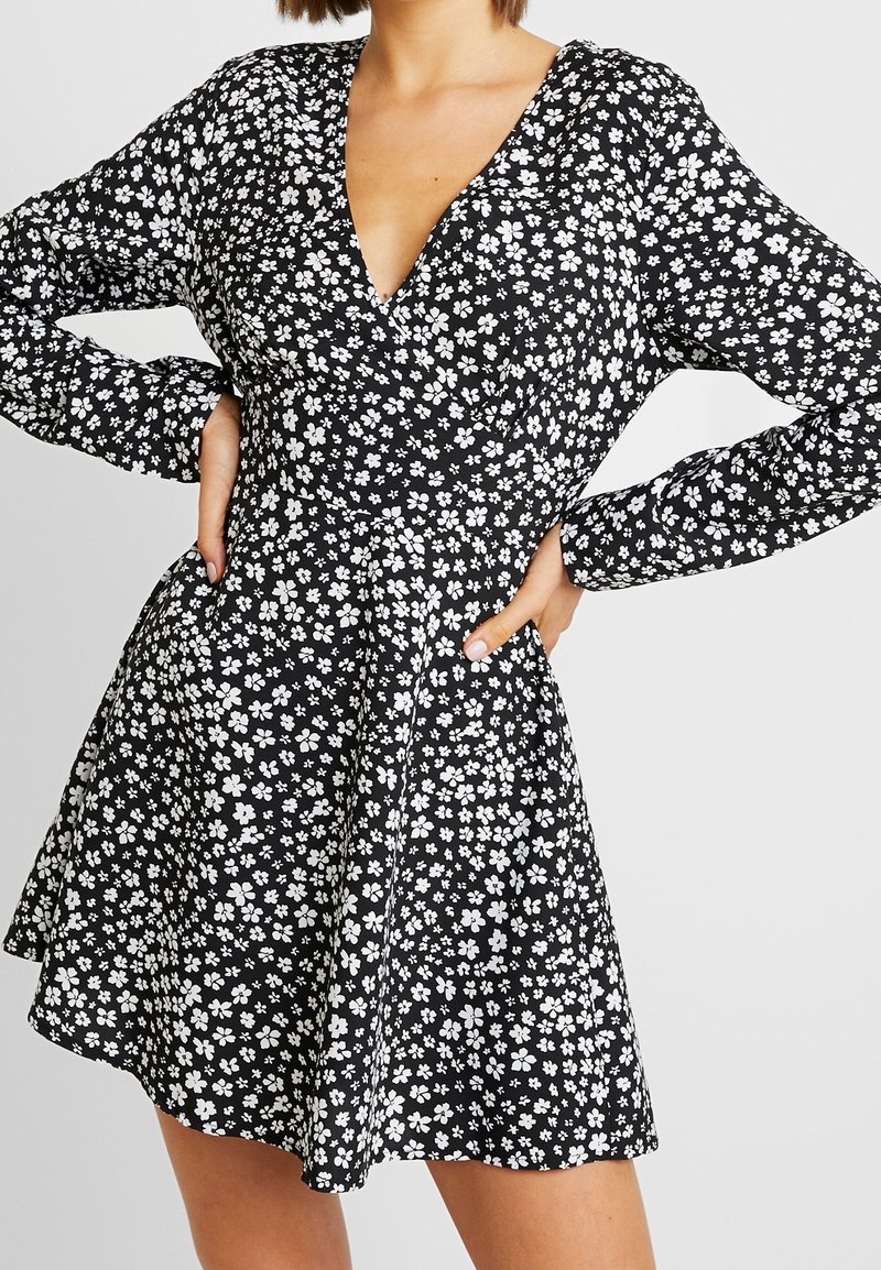 Femme portant une robe noire à manches longues avec un motif floral blanc, les mains sur les hanches, debout devant un fond uni.