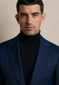 Granatowa marynarka w delikatny wzór w kratę, zestawiona z czarnym golfem. Strukturalne klapy i dopasowany krój podkreślają design.