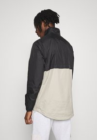 Giacca a vento nera e beige con colletto alto, polsini elasticizzati e design a mezza zip. Tessuto morbido, leggero.