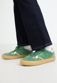 Zapatillas de ante verde con acentos en rosa y amarillo, suelas beige y una superficie texturizada. Diseño con cordones, combinadas con jeans de mezclilla oscura.