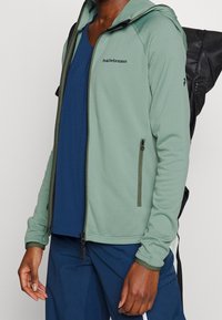 Veste zippée verte avec capuche et poches zippées, en matière extensible, avec logo brodé et accents foncés.