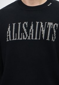 Gros plan d'un pull en maille noir avec un motif de point de croix blanc formant le mot "ALLSAINTS" sur la poitrine et trois petites croix près du col.