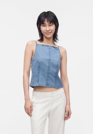 Mujer joven con el pelo corto y negro, vistiendo una blusa de mezclilla azul sin mangas y pantalones blancos, de pie frente a un fondo claro y liso.