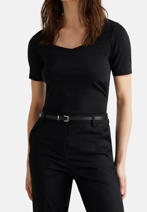 Kobieta w dopasowanym czarnym topie z krótkim rękawem i dekoltem w serek, wpuszczonym w czarne spodnie z wysokim stanem, przewiązane cienkim czarnym paskiem.