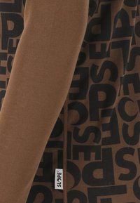 Brauner Stoff mit großen schwarzen, überlappenden Buchstaben und einem kleinen weißen Etikett mit der Aufschrift "SLOPE", das in eine Naht eingenäht ist.