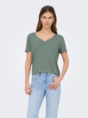 Young woman with long brown hair wears green short-sleeve V-neck top with lace trim and light blue high-waisted jeans, standing against plain background.