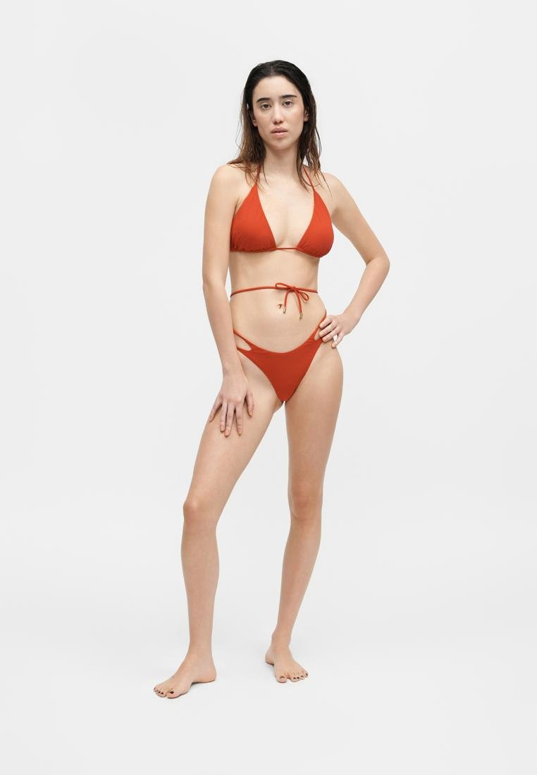 Jeune femme debout pieds nus portant un bikini couleur rouille avec un haut triangle et un bas découpé sur un fond blanc uni.