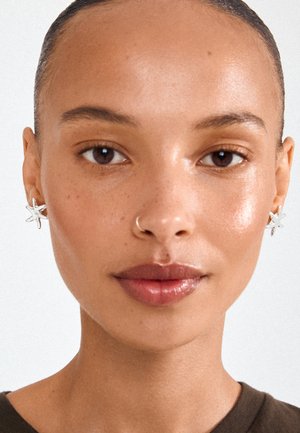 Vrouw met een gladde bruine huid, zilveren stervormige oorbellen en een klein neusringetje, met haar naar achteren en neutrale make-up.