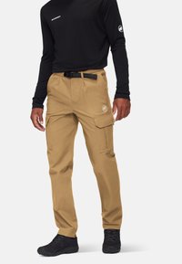 Mężczyzna w beżowych spodniach cargo z kieszeniami na klapki i czarnym paskiem, w połączeniu z czarną koszulą z długim rękawem i czarnymi butami, stojący na białym tle.