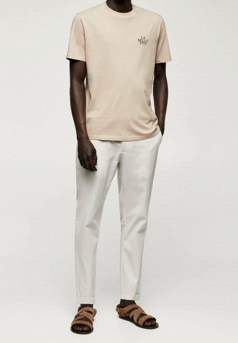 Homme portant un t-shirt beige avec l'inscription « La Bella Vita », pantalon blanc et sandales marron, debout avec une main dans la poche.