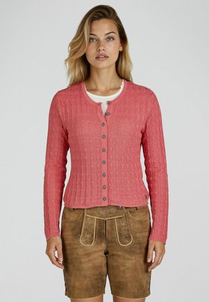 Vrouw met een koraalroze kabelgebreide vest met knopen over een wit topje en traditionele bruine suède leren shorts.